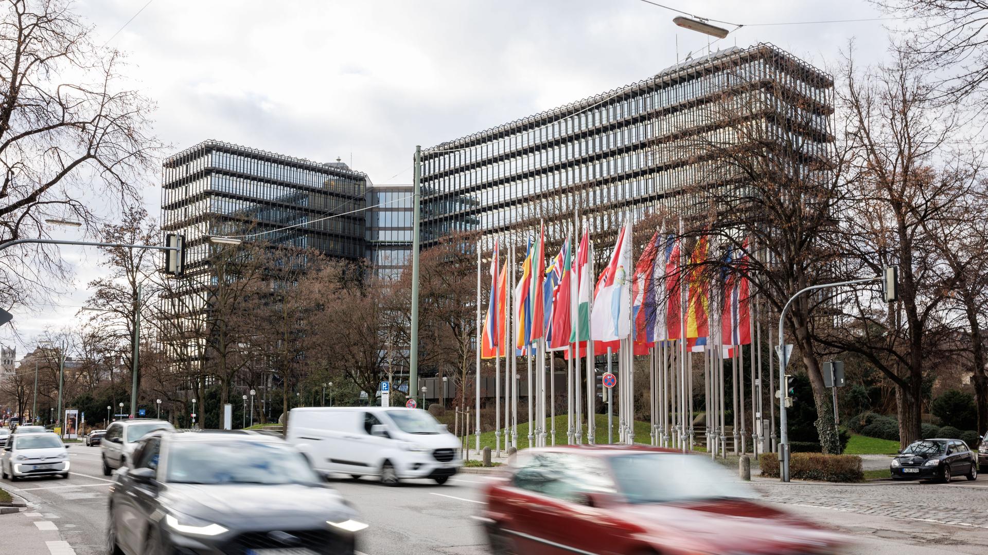 Blick von außen auf den Gebäudekomplex aus Glas und Stahl. Davor die Flaggen der EU-Staaten. Über eine breite Straße im Vordergrund fahren viele Autos. 