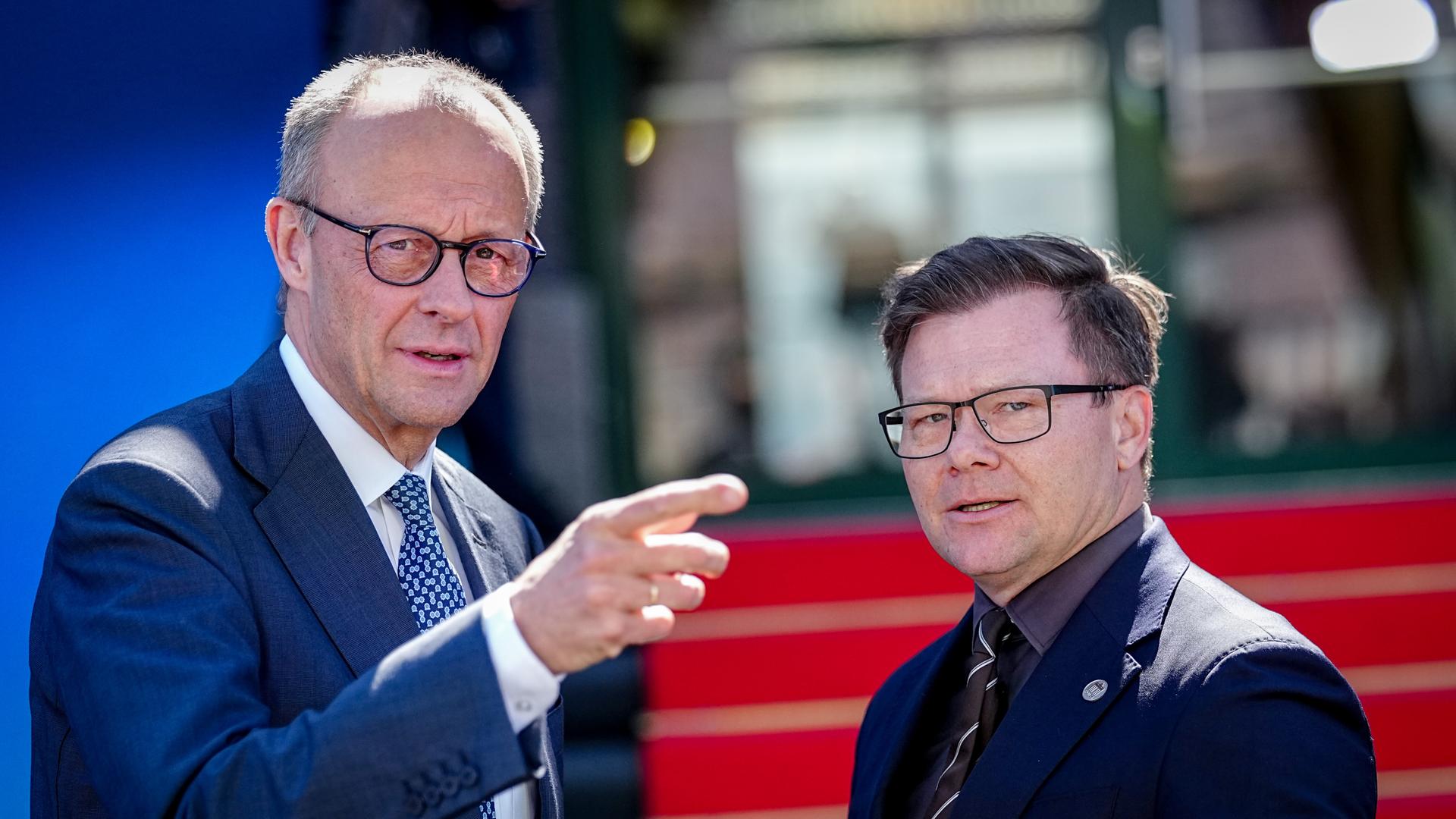 Friedrich Merz und Carsten Schneider stehen nebeneinander im Freien. Merz, links im Bild, trägt einen blauen Anzug mit gemusterter Krawatte, spricht und zeigt mit ausgestrecktem Arm nach rechts aus dem Bild.