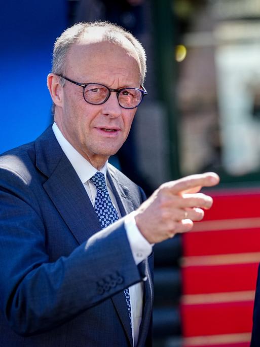Friedrich Merz und Carsten Schneider stehen nebeneinander im Freien. Merz, links im Bild, trägt einen blauen Anzug mit gemusterter Krawatte, spricht und zeigt mit ausgestrecktem Arm nach rechts aus dem Bild.