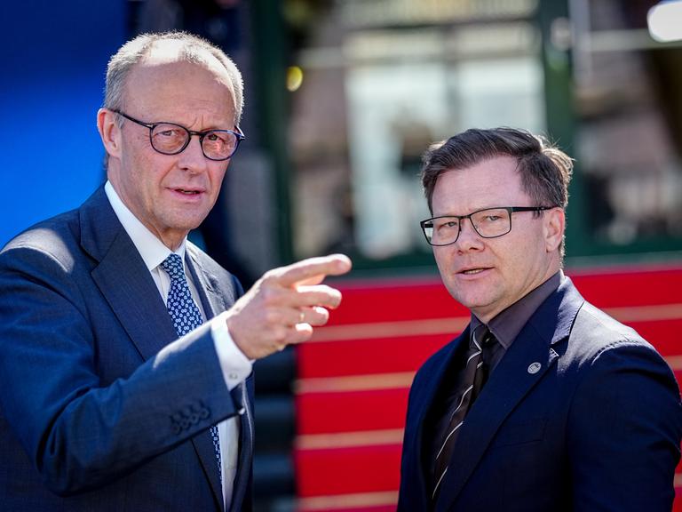 Friedrich Merz und Carsten Schneider stehen nebeneinander im Freien. Merz, links im Bild, trägt einen blauen Anzug mit gemusterter Krawatte, spricht und zeigt mit ausgestrecktem Arm nach rechts aus dem Bild.