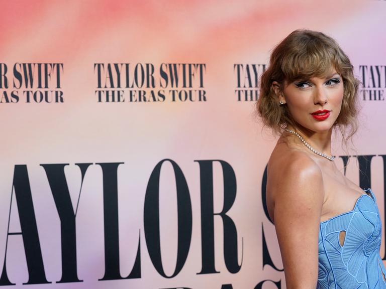 Taylor Swift guckt ganz rechts im Bilder über ihre Schulter. Sie steht vor einem Plakat mit der Aufschrift: "Taylor Swift: The Eras Tour".