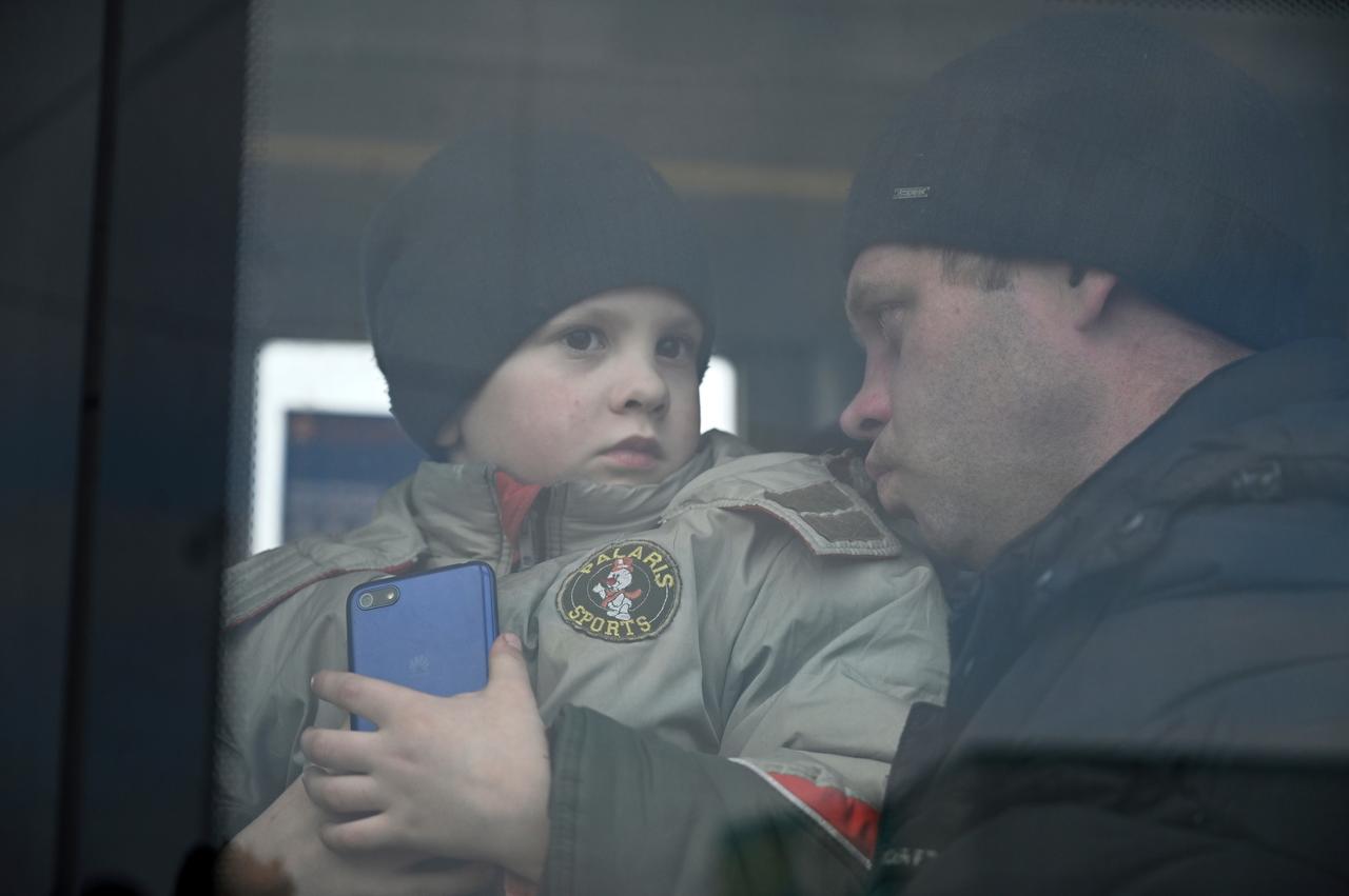 Blick von außen durch die Scheibe. Der Junge schaut traurig nach draußen, sein Vater scheut ihn besorgt an. Beide tragen schwarze Wollmützen. Der Junge hält ein Handy in der Hand.  