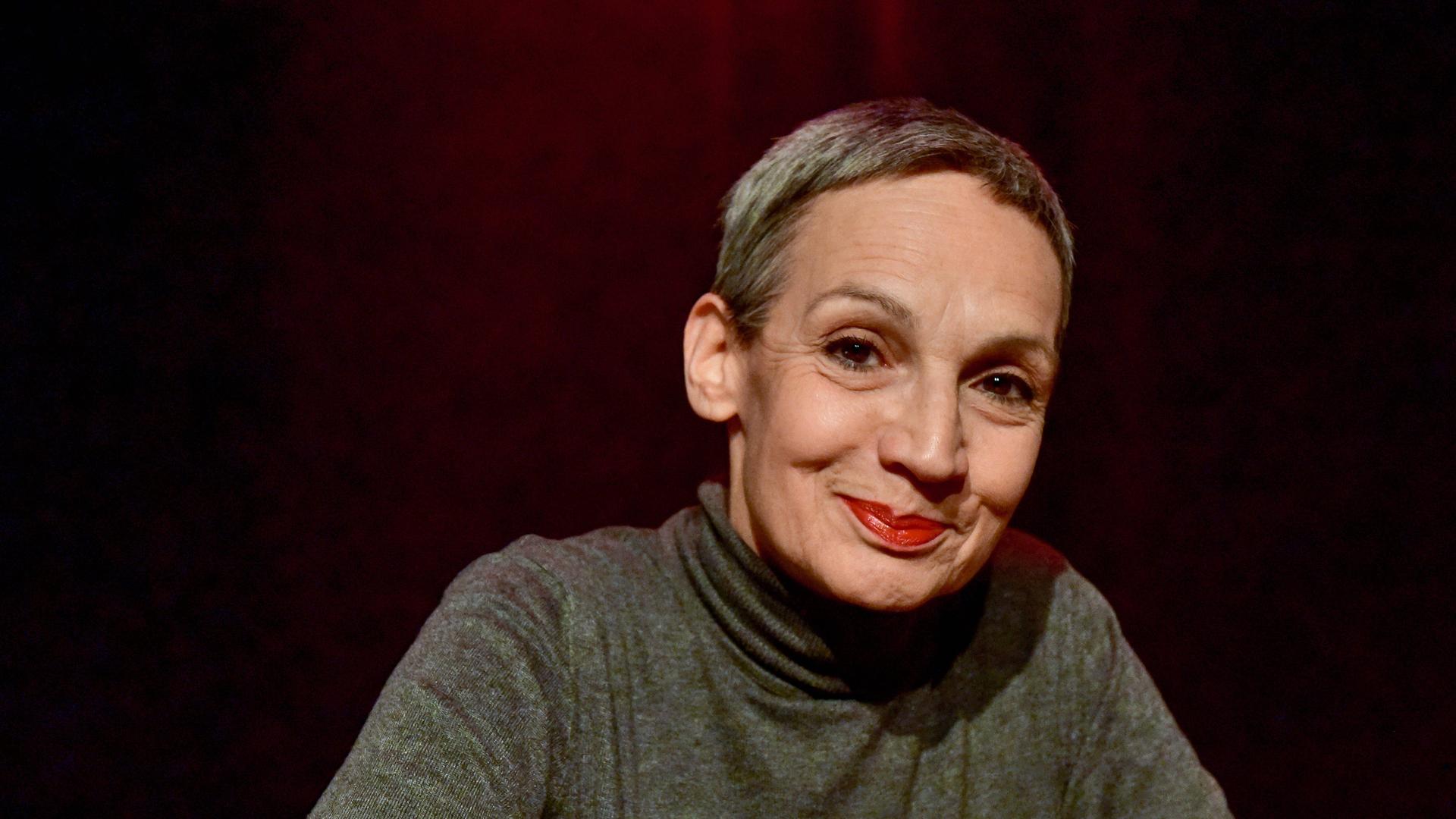 Marion Brasch hat graue Haare und trägt eine Kurzhaarfrisur. Auf dem Foto trägt sie Lippenstift und einen grauen Pullover. Der Hintergrund ist dunkel.
