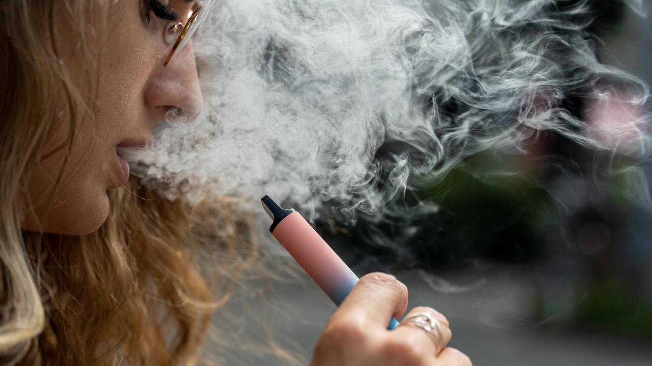 Eine junge Frau hält eine Vape, eine E-Zigarette, in der Hand. Aus ihrem Mund kommt Rauch heraus.