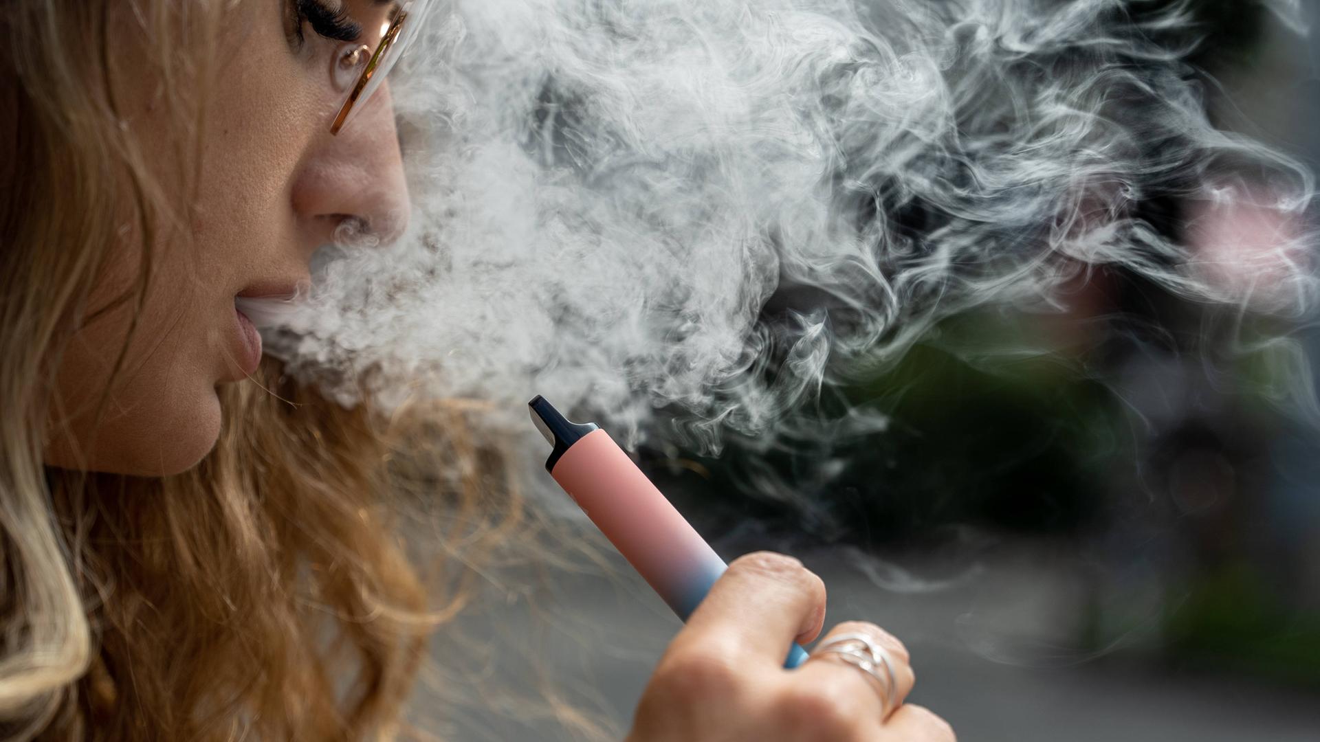 Eine Frau hält eine Vape, eine E-Zigarette, in der Hand. Aus ihrem Mund kommt Rauch heraus.