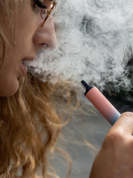Eine Frau hält eine Vape, eine E-Zigarette, in der Hand. Aus ihrem Mund kommt Rauch heraus.