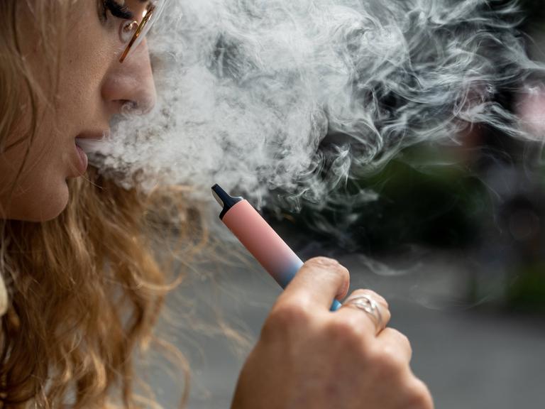 Eine Frau hält eine Vape, eine E-Zigarette, in der Hand. Aus ihrem Mund kommt Rauch heraus.