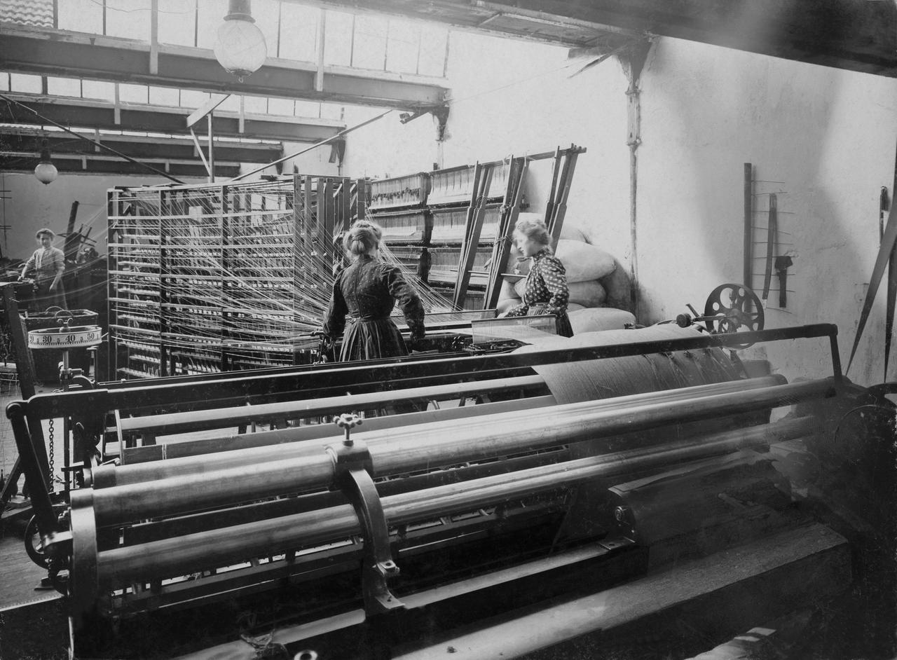  Frauen stehen an einer Maschine, aus der gesponnenes Garn kommt. Um 1910.