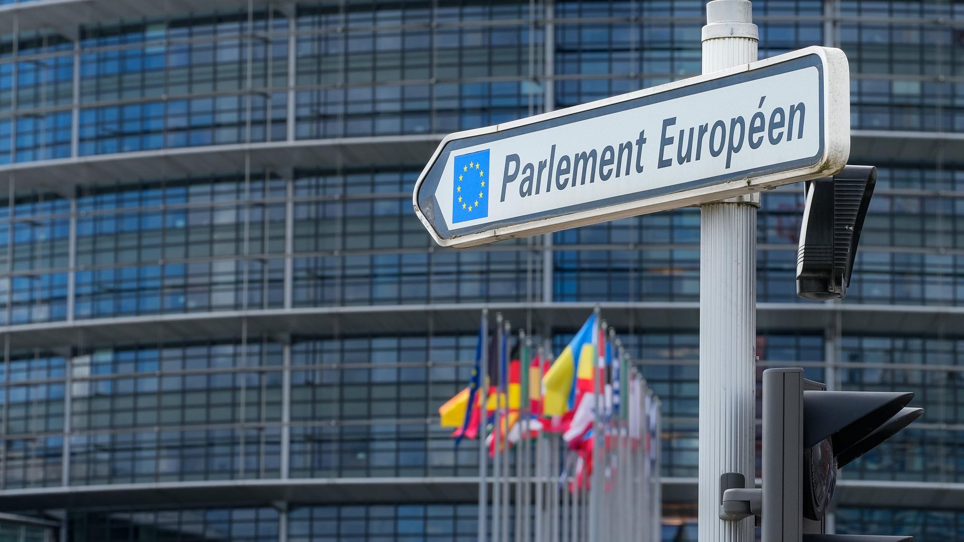 Hinter einem Wegweiser mit der Aufschrift "Parlement Européen" sind die Flaggen der Mitgliedssaaten der EU und der Sitz des EU-Parlaments in Straßburg zu sehen. 