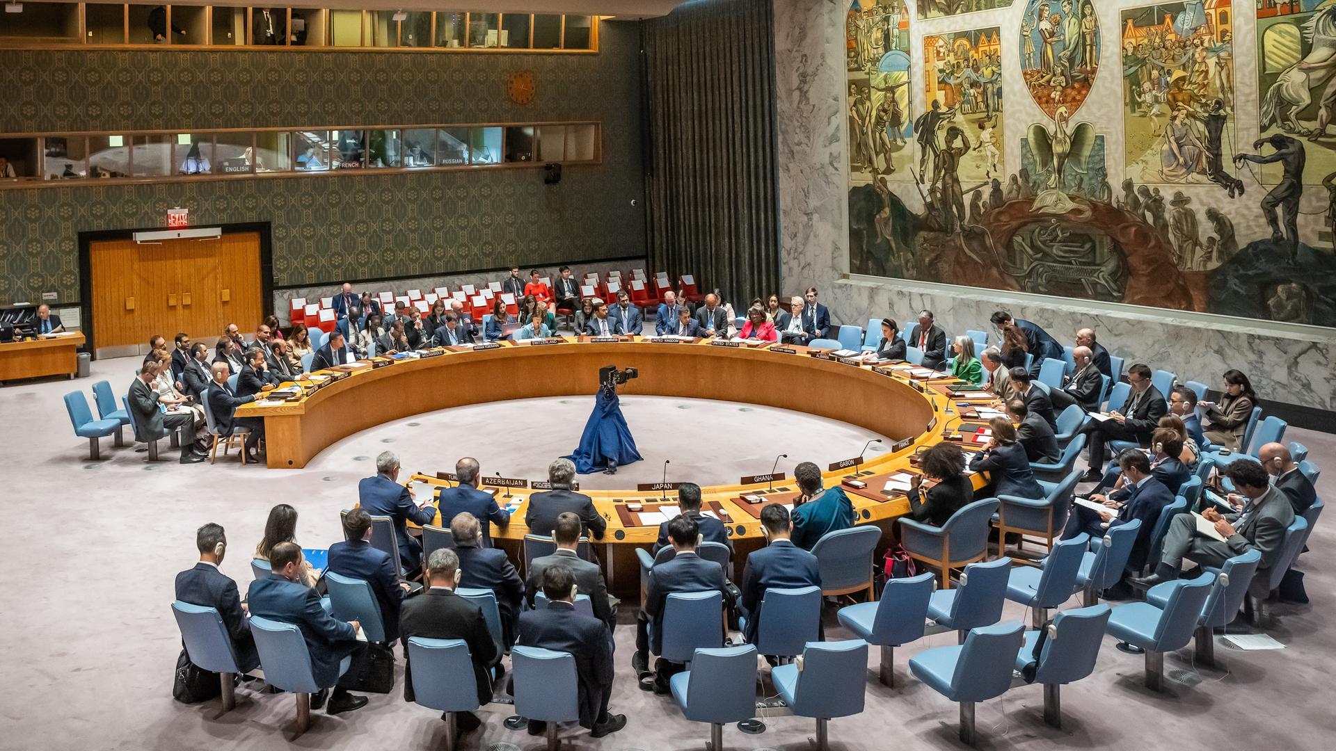 Näkymä turvallisuusneuvoston saliin, jossa kaiutinpöytä on asetettu ympyrään sinisillä tuoleilla ja taidokkaalla seinämaalauksella.