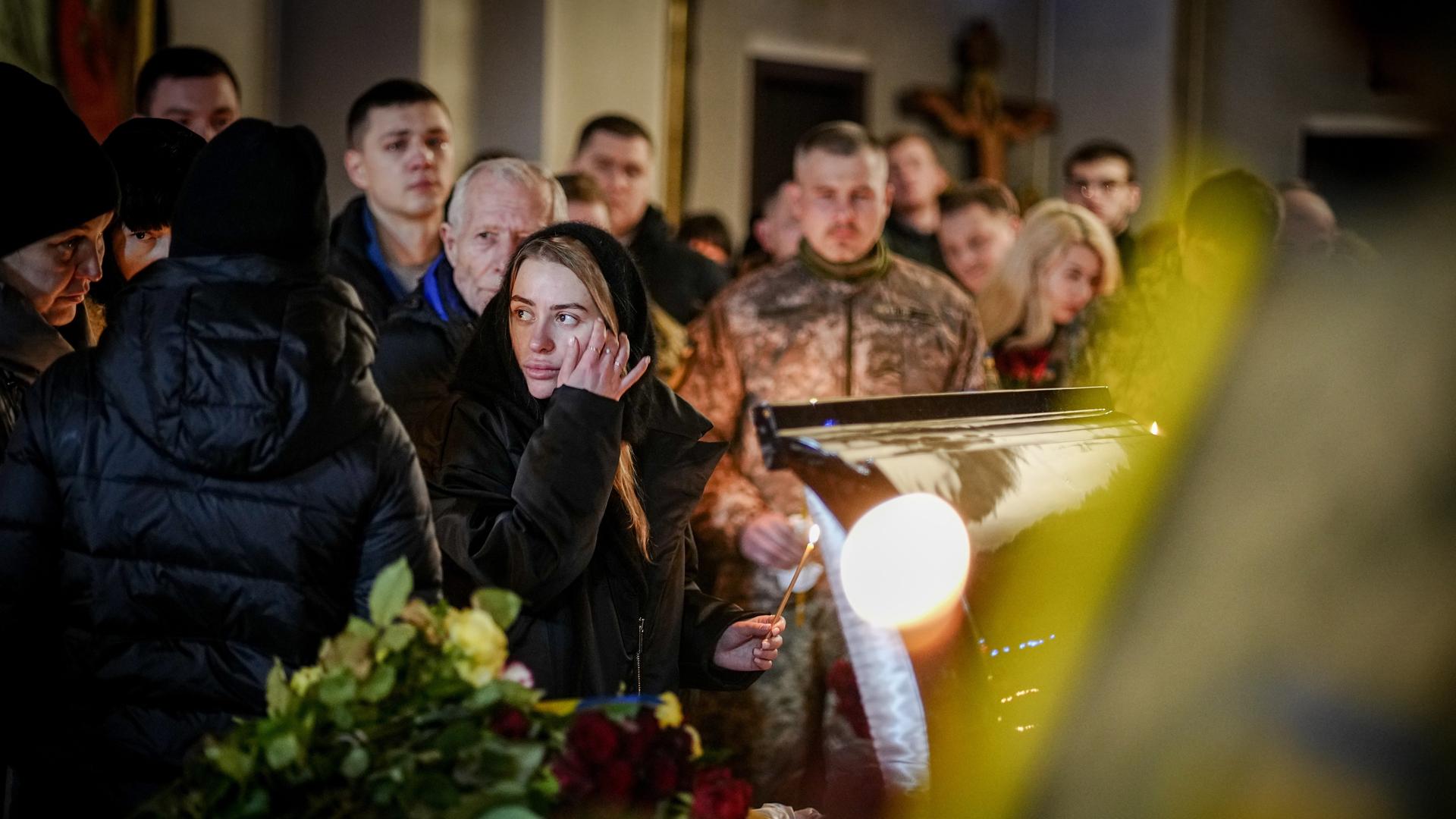 Eine Frau mit Kerze in der Hand steht vor dem Sarg eines getöteten Soldaten. Um sie herum weitere Trauernde.