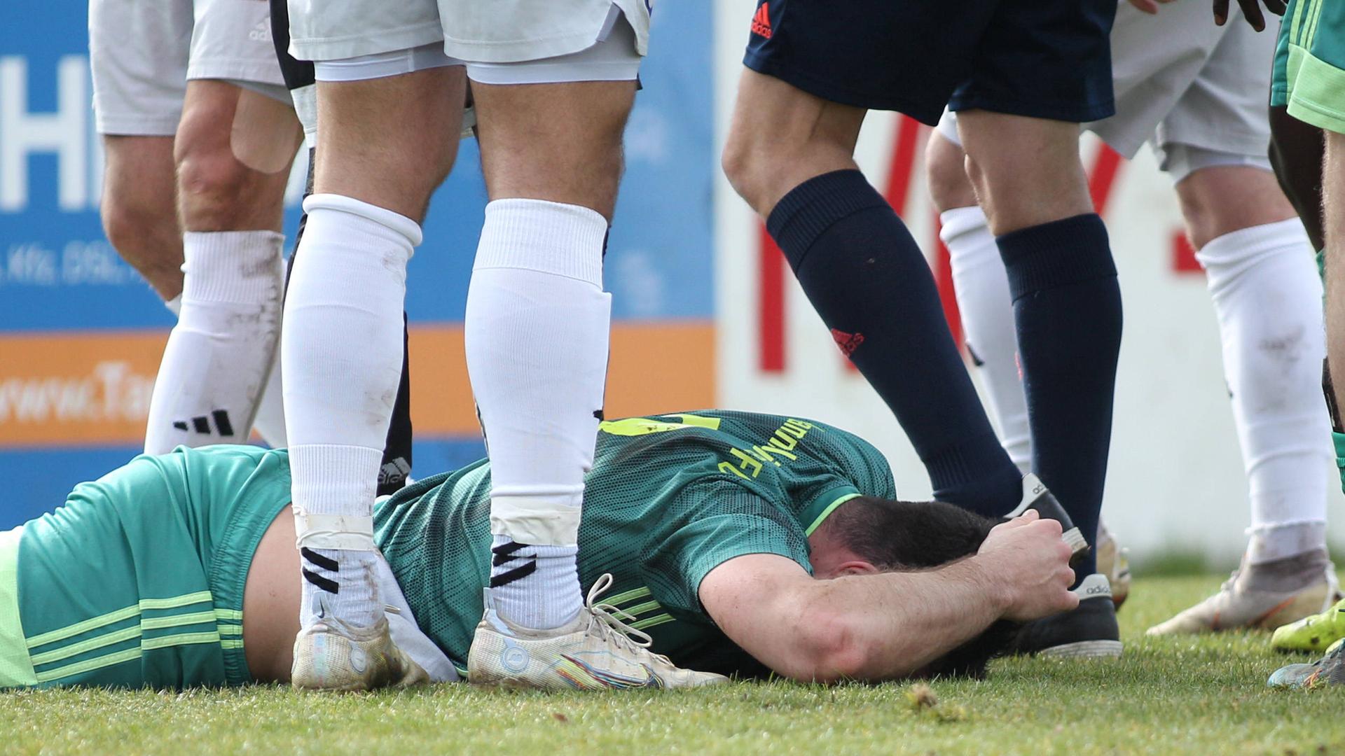 Ein Fußballspieler liegt auf dem Boden und hält sich den Kopf. Daneben sind Beine weiterer Spieler und des Schiedsrichters.
