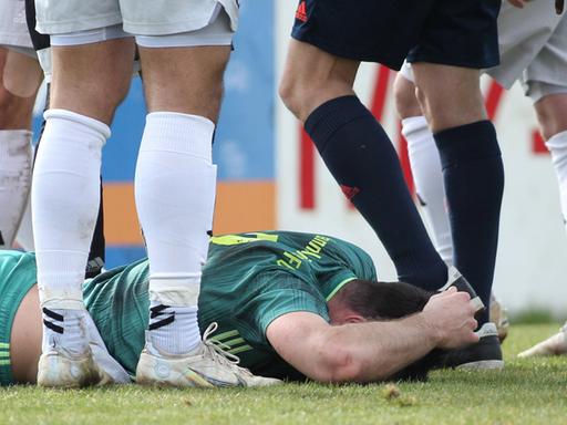 Ein Fußballspieler liegt auf dem Boden und hält sich den Kopf. Daneben sind Beine weiterer Spieler und des Schiedsrichters.