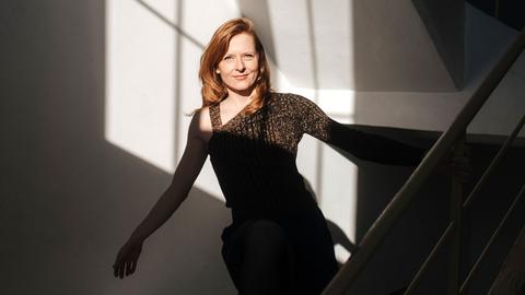 Eine Frau mit rötlichen Haaren steht mit ausgestreckten Armen auf einer Treppe. Ihr Körper liegt teils im Schatten, teils im hellen Licht, das aus einem gegenüberliegenden, nicht sichtbaren Fenster auf sie fällt. Sie trägt ein Glitzeroberteil.