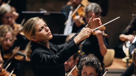 Blick auf die Dirigentin mit erhobenen Händen vor ihrem Orchester stehend.