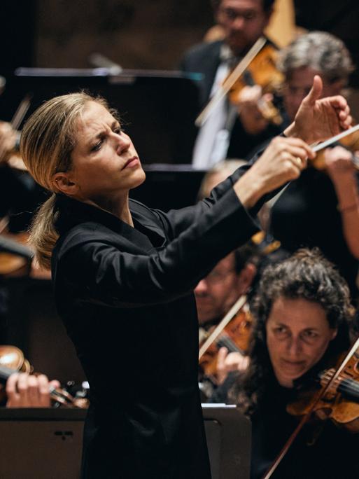 Blick auf die Dirigentin mit erhobenen Händen vor ihrem Orchester stehend.