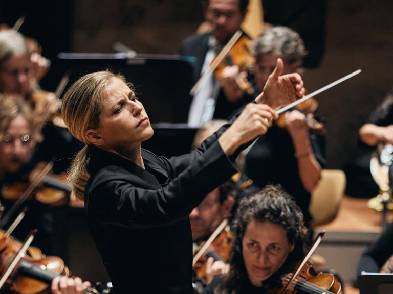 Blick auf die Dirigentin mit erhobenen Händen vor ihrem Orchester stehend.