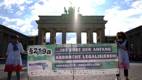 Vor dem Brandenburger Tor halten zwei Frauen ein Plakat hoch, auf dem steht: Paragraf 219a ist erst der Anfang, Abbrüche legalisieren.