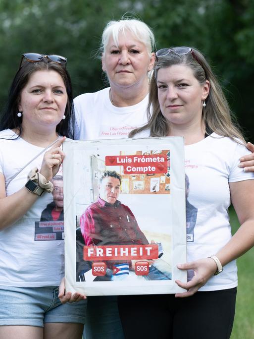 Maria (l) und Janie (r) Frometa Compte, Töchter des inhaftierten Deutsch-Kubaners Luis Frómeta Compte, und Silke Frometa Compte (M), Ehefrau, halten ein Foto von Luis Frometa Compte.