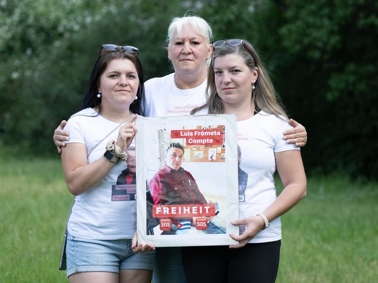 Maria (l) und Janie (r) Frometa Compte, Töchter des inhaftierten Deutsch-Kubaners Luis Frómeta Compte, und Silke Frometa Compte (M), Ehefrau, halten ein Foto von Luis Frometa Compte.