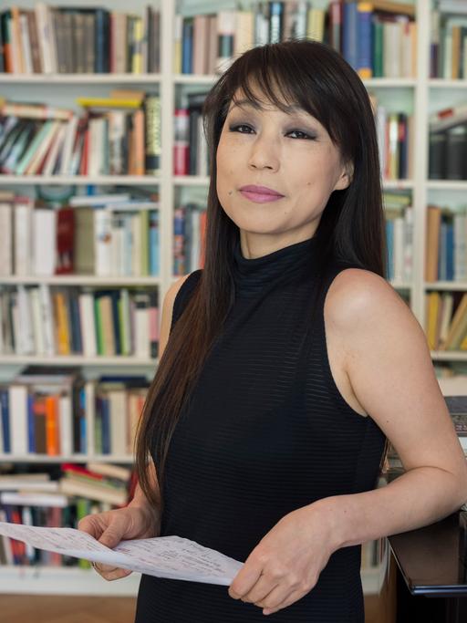 Eine Frau mit langem dunklem Haar lehnt sich an einen Flügel. Im Hintergrund ist ein Bücherregal zu sehen.
