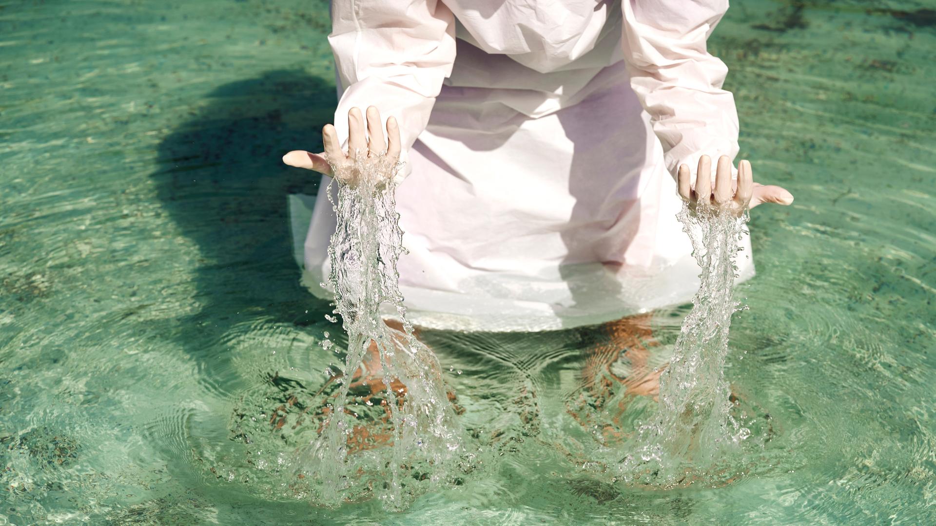 Eine Person in einem weißen Gewand steht im Wasser und hebt die Hände aus dem Wasser. (Symbolbild)