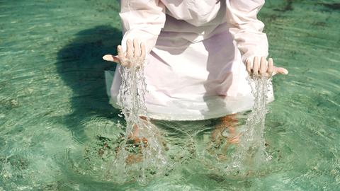 Eine Person in einem weißen Gewand steht im Wasser und hebt die Hände aus dem Wasser. (Symbolbild)