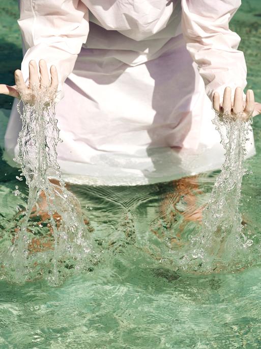 Eine Person in einem weißen Gewand steht im Wasser und hebt die Hände aus dem Wasser. (Symbolbild)