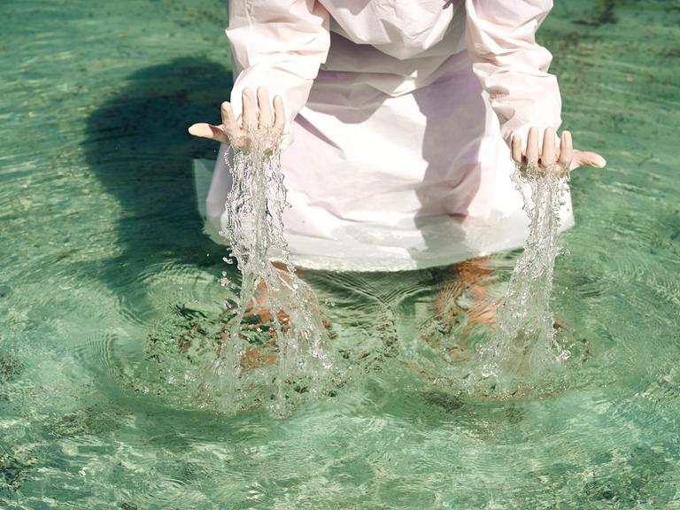 Eine Person in einem weißen Gewand steht im Wasser und hebt die Hände aus dem Wasser. (Symbolbild)