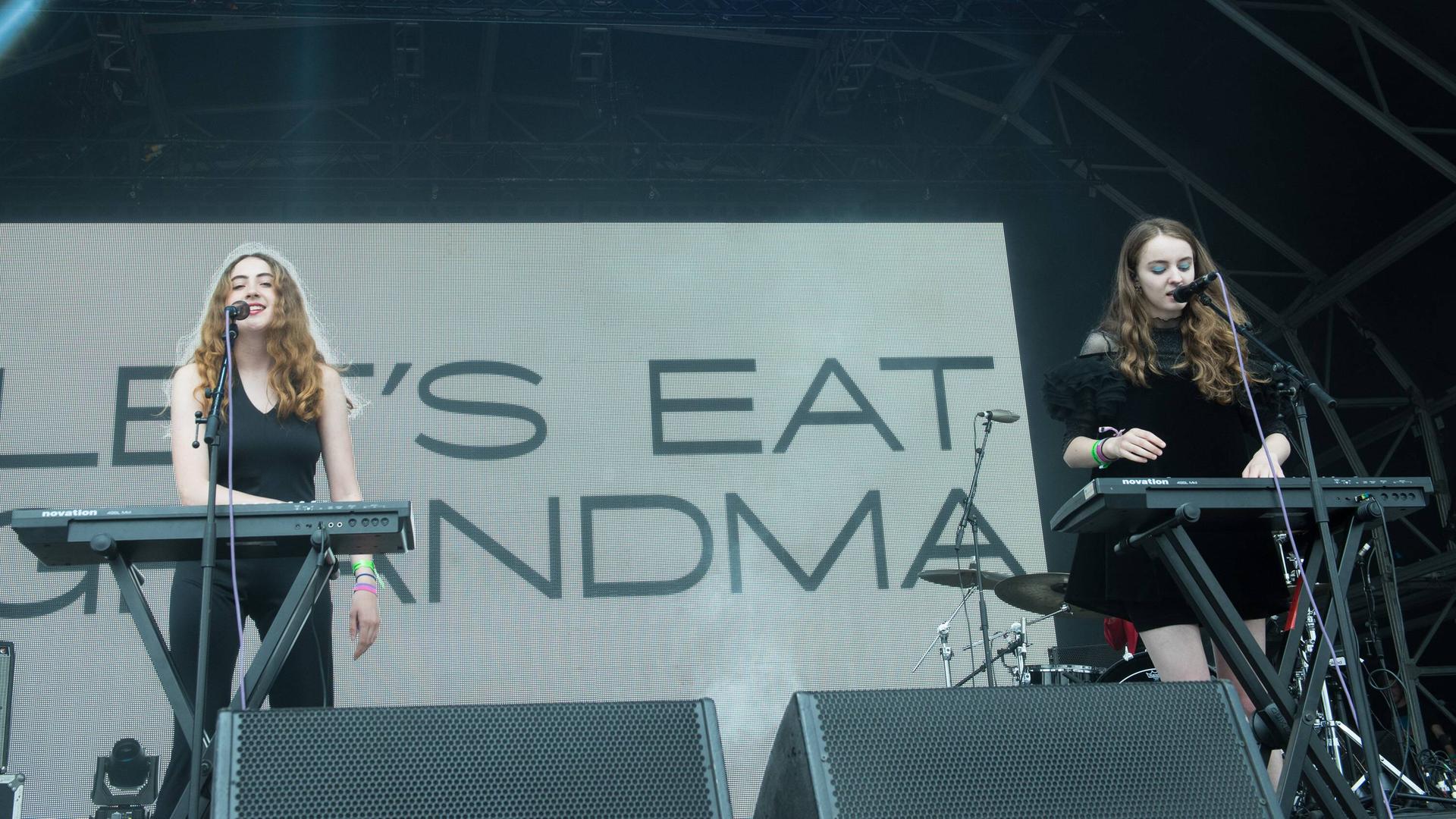 Zwei Frauen an Keyboards stehen vor einer Leinwand auf der "Let's eat grandma" steht.