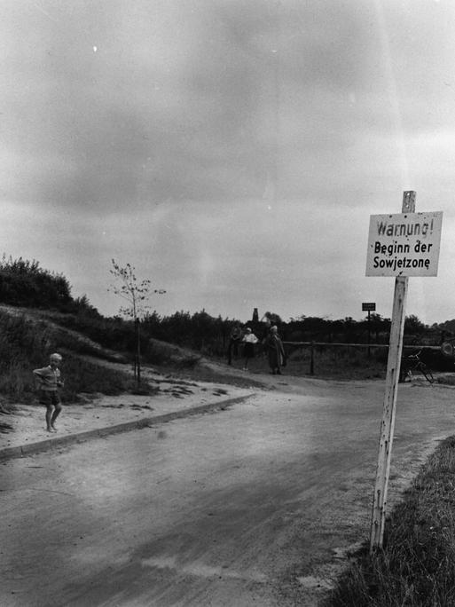 Warnschild "Beginn der Sowjetzone" an einer ländlichen Straße mit Bushaltestelle. Kinder spielen.