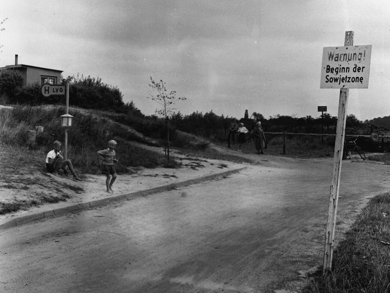 Warnschild "Beginn der Sowjetzone" an einer ländlichen Straße mit Bushaltestelle. Kinder spielen.
