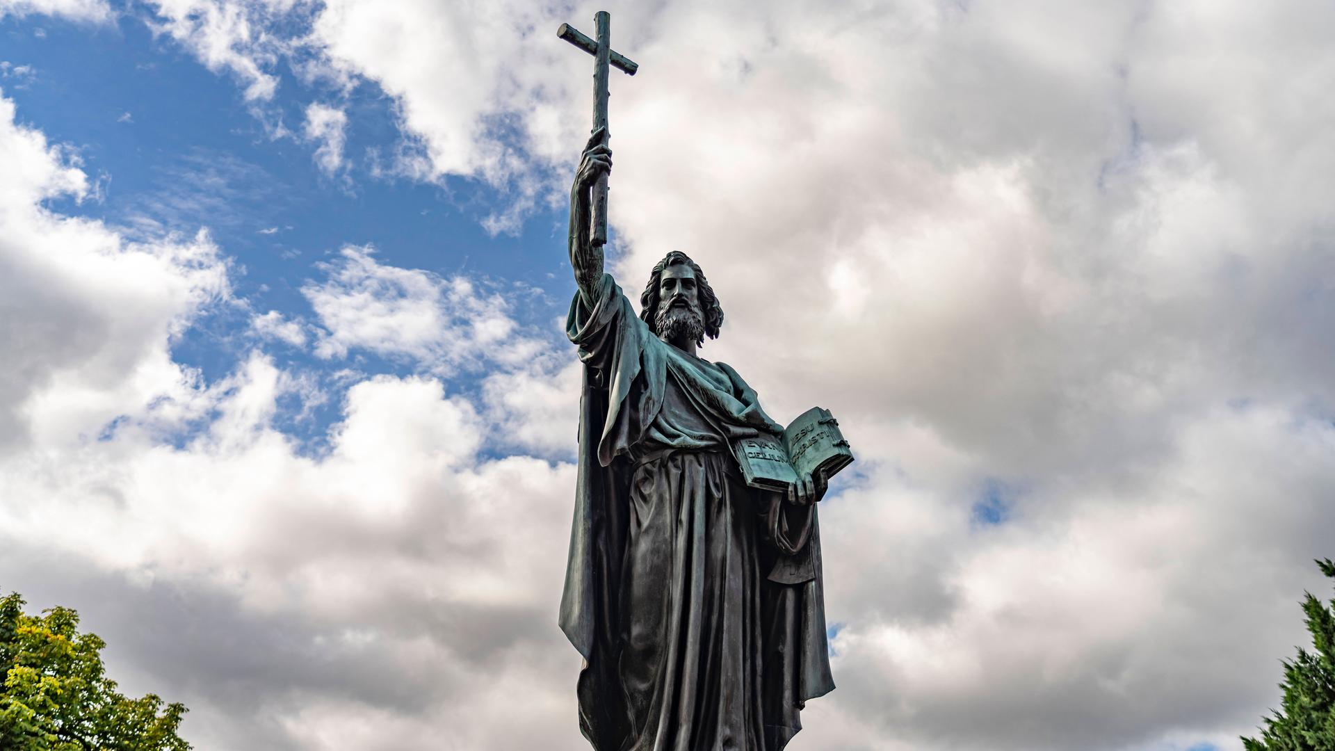 Die metallene Figur eines männlichen Heiligen steht in der Landschaft herum. Der Rechte Arm ist in die Höhe gereckt – mit einem Kreuz in der Hand. Die Rechte hält lässig ein geöffnetes Buch. Das Gesicht ist stark behaart.