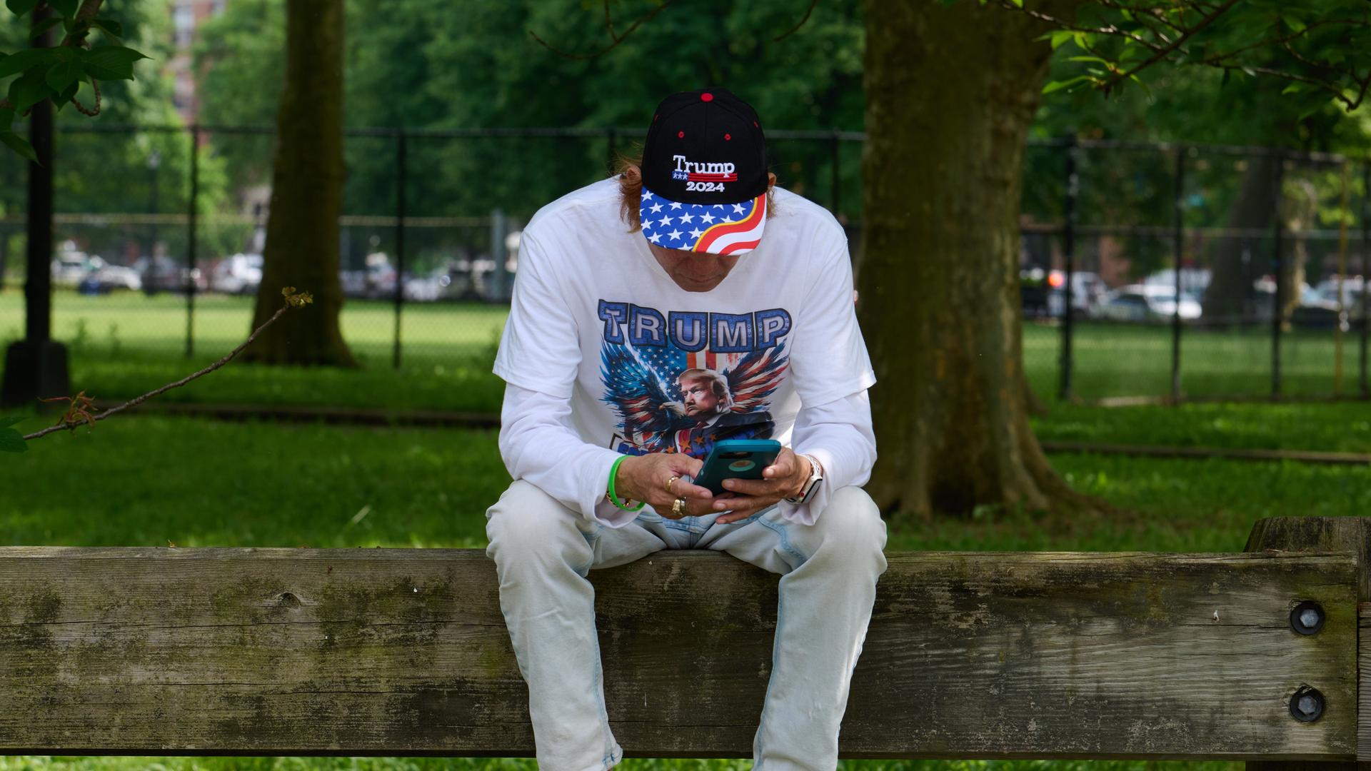 Mann mit Trump-Shirt und Basecap sitzt auf Rückenlehne einer Bank und blickt aufs Smartphone (25.5.2024). Mann mit Trump-Shirt und Basecap sitzt auf Rückenlehne einer Bank und blickt aufs Smartphone (25.5.2024).