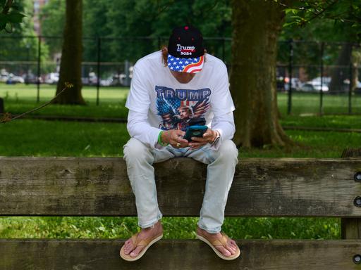 Mann mit Trump-Shirt und Basecap sitzt auf Rückenlehne einer Bank und blickt aufs Smartphone (25.5.2024). Mann mit Trump-Shirt und Basecap sitzt auf Rückenlehne einer Bank und blickt aufs Smartphone (25.5.2024).