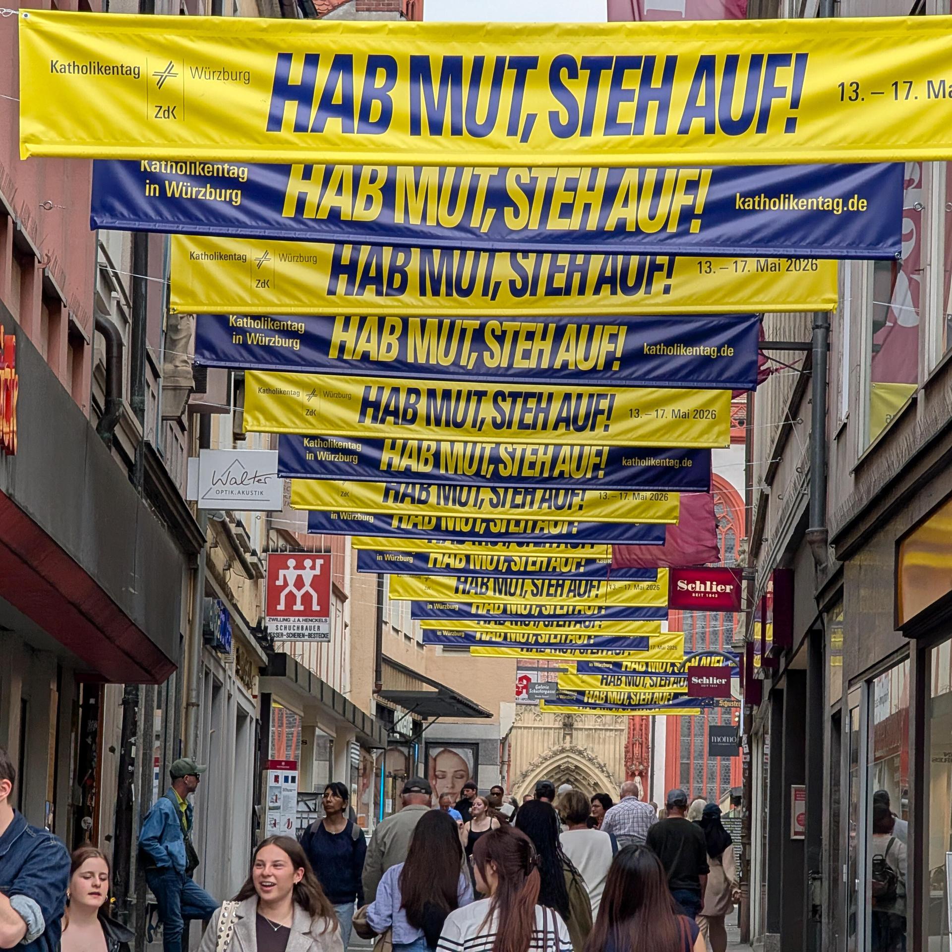 Blaue und gelbe Banner für den Kirchentag sind in der Fußgängerzone von Würzburg angebracht. Darauf steht "Hab Mut, steh auf!". Darunter gehen mehrere Fußgänger.