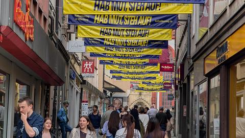 Blaue und gelbe Banner für den Kirchentag sind in der Fußgängerzone von Würzburg angebracht. Darauf steht "Hab Mut, steh auf!". Darunter gehen mehrere Fußgänger.