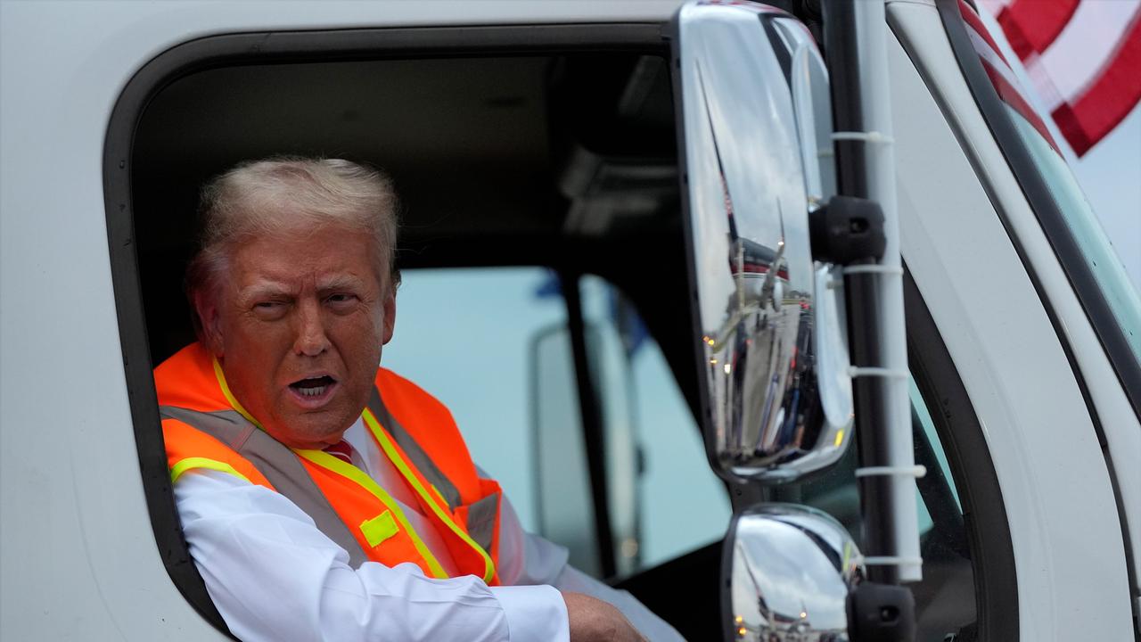 Trump trägt eine orange-gelbe Weste und lehnt sich aus dem Fenster des Müllwagen heraus. 