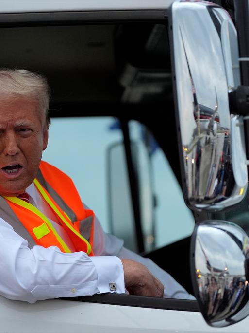 Trump trägt eine orange-gelbe Weste und lehnt sich aus dem Fenster des Müllwagen heraus. 