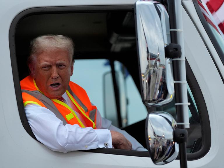 Trump trägt eine orange-gelbe Weste und lehnt sich aus dem Fenster des Müllwagen heraus. 