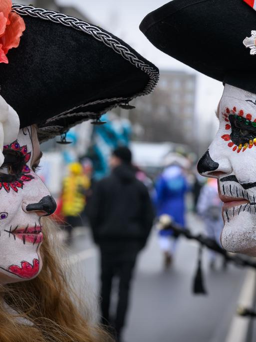 Zwei Teilnehmer am Karnevalsumzug "Zug der fröhlichen Leute" schauen sich in die Augen. Im Hintergrund sind einige andere Menschen zu sehen, teils in Kostümen.