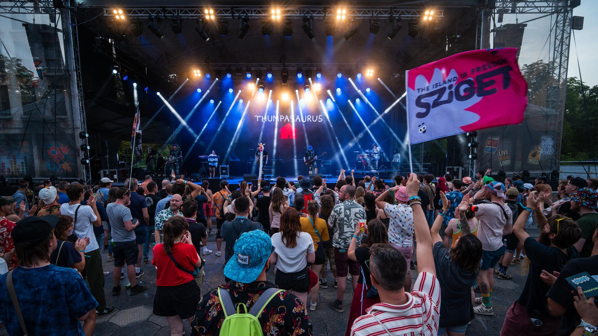 Menschen stehen vor einer Bühne, eine Person hält eine Fahne mit der Aufschrift "Sziget"