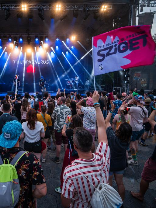 Menschen stehen vor einer Bühne, eine Person hält eine Fahne mit der Aufschrift "Sziget" Menschen stehen vor einer Bühne, eine Person hält eine Fahne mit der Aufschrift "Sziget"