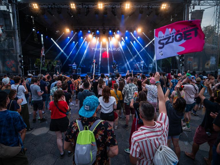 Menschen stehen vor einer Bühne, eine Person hält eine Fahne mit der Aufschrift "Sziget" Menschen stehen vor einer Bühne, eine Person hält eine Fahne mit der Aufschrift "Sziget"