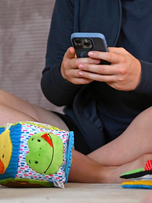 Ein Baby krabbelt von seiner Mutter weg, die auf dem Boden sitzt und auf ihr Handy schaut. 