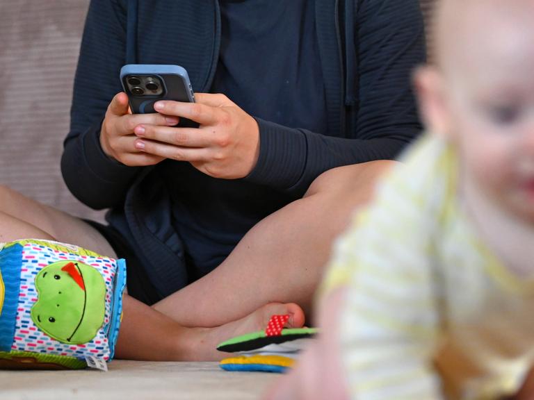 Ein Baby krabbelt von seiner Mutter weg, die auf dem Boden sitzt und auf ihr Handy schaut. 