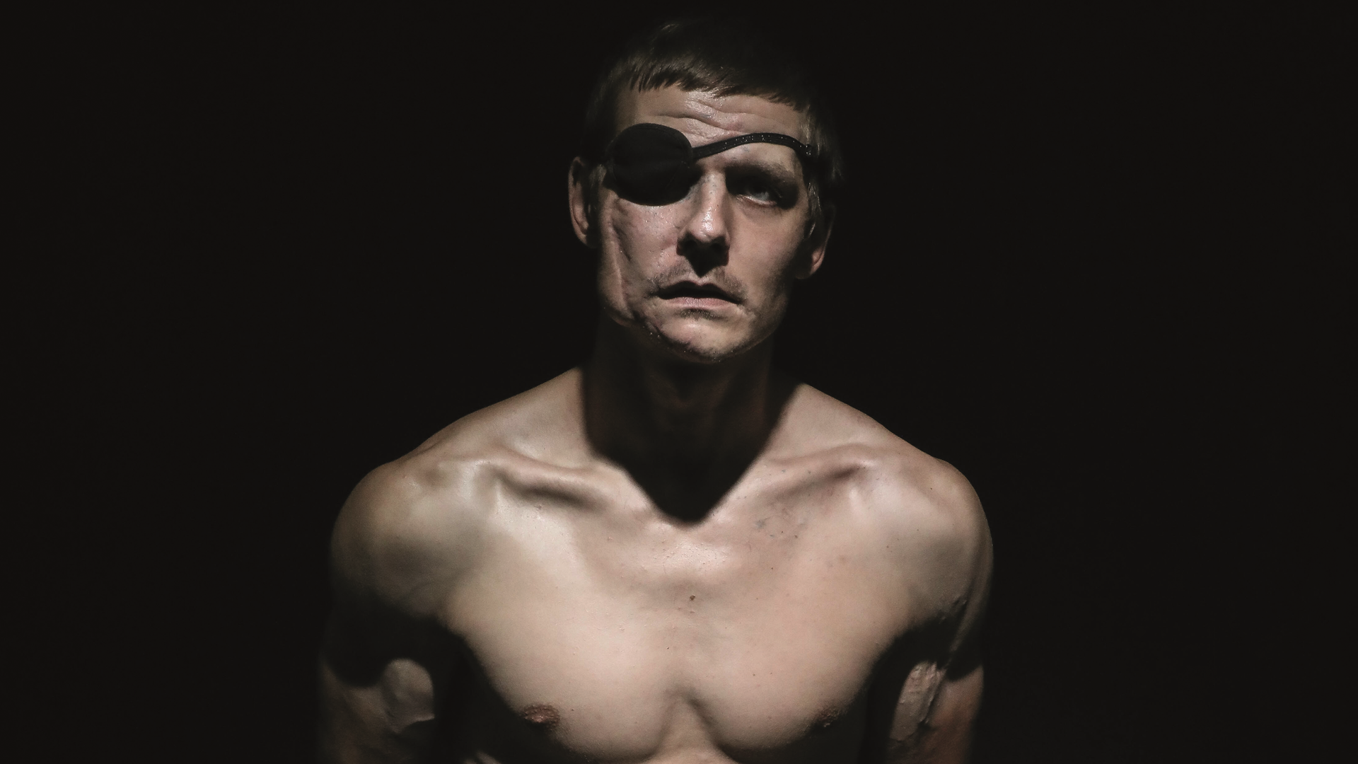 Der verwundete ukrainische Soldat Wolodymyr Melnik sitzt mit nacktem Oberkörper in einem dunklen Fotostudio. Er trägt eine Augenklappe.