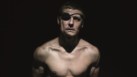 Der verwundete ukrainische Soldat Wolodymyr Melnik sitzt mit nacktem Oberkörper in einem dunklen Fotostudio. Er trägt eine Augenklappe.