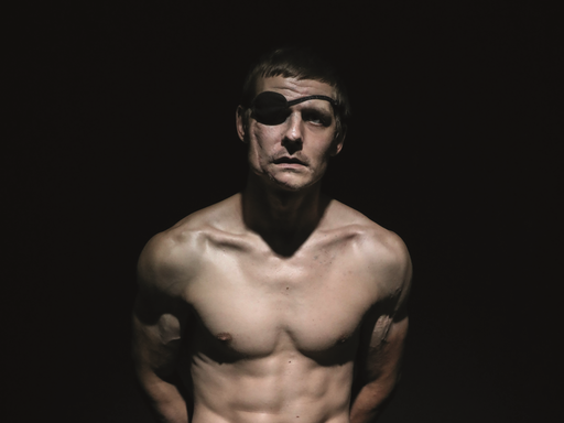 Der verwundete ukrainische Soldat Wolodymyr Melnik sitzt mit nacktem Oberkörper in einem dunklen Fotostudio. Er trägt eine Augenklappe.