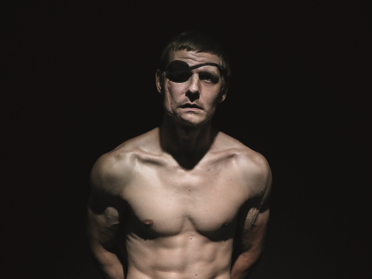 Der verwundete ukrainische Soldat Wolodymyr Melnik sitzt mit nacktem Oberkörper in einem dunklen Fotostudio. Er trägt eine Augenklappe.